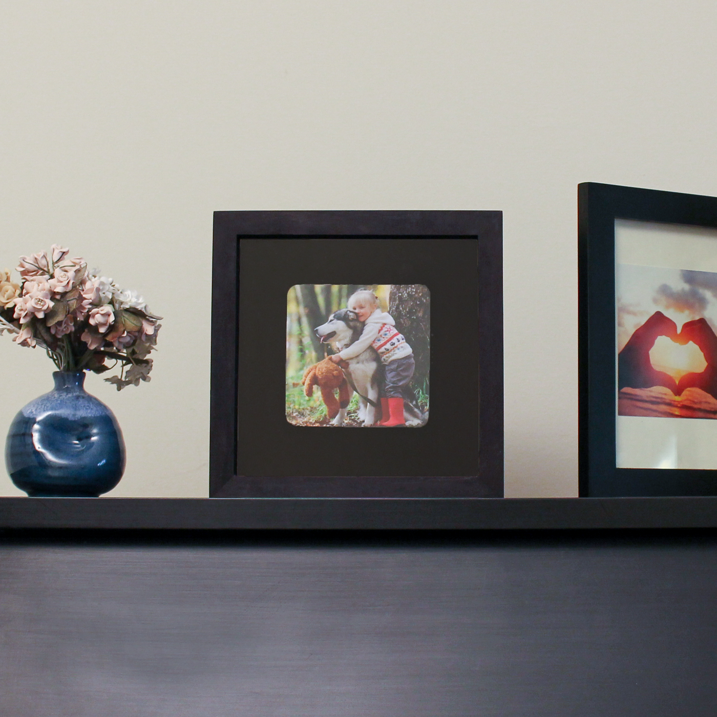 An arrangement of home decor items: A blue vase with pink flowers with green stems, a black picture frame along with a picture of a young firl and a dog, and a portion of another picture that shows hands in the shape of a heart. 